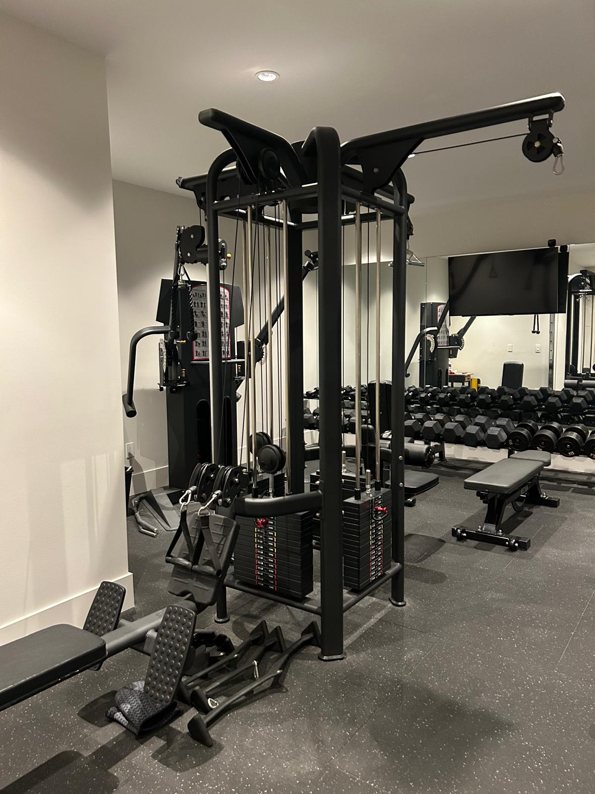 Dual-stack functional cable trainer with adjustable benches, dumbbell rack, mounted TV, and mirrored wall in the Abbotsford home gym build.