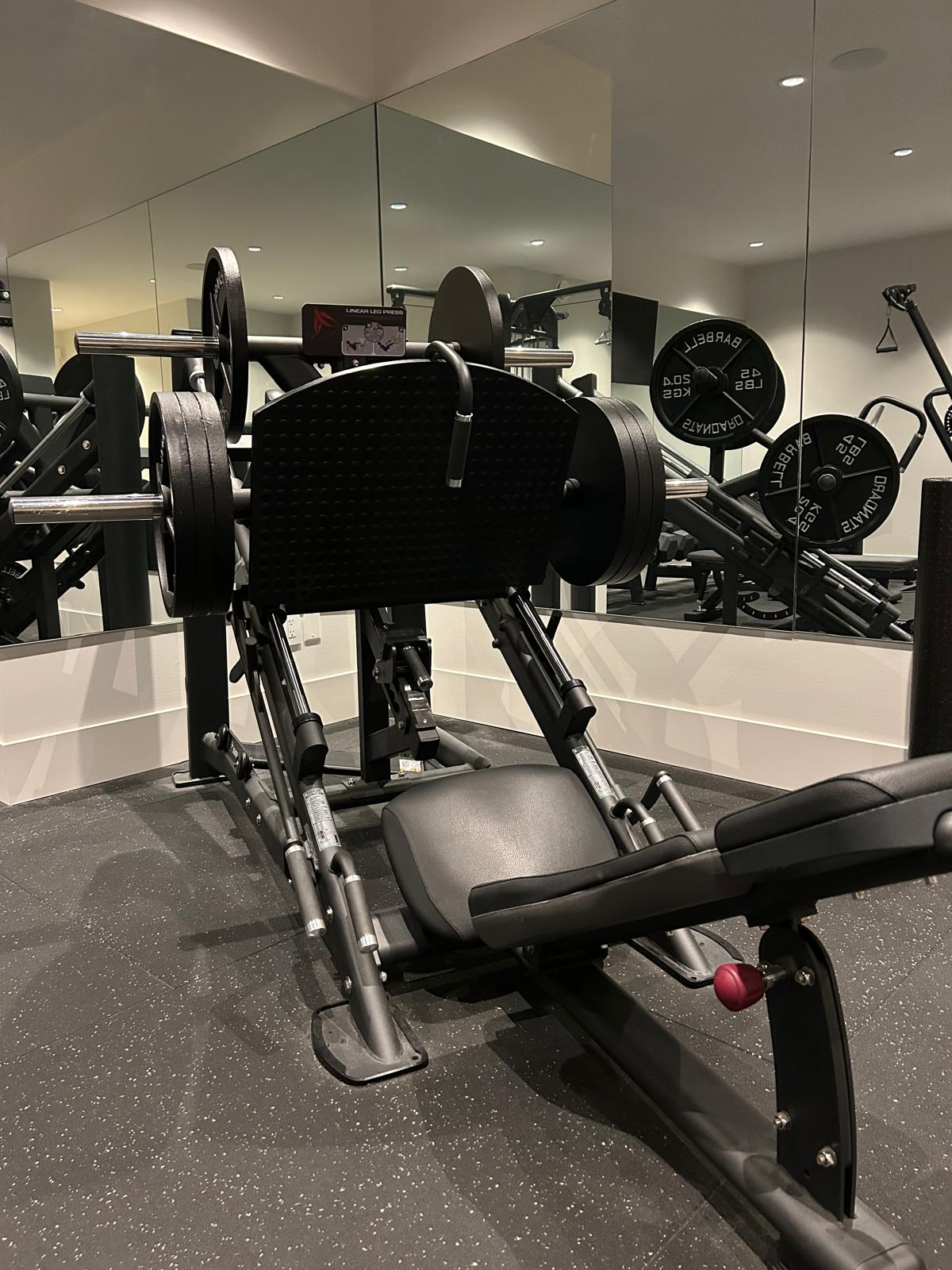 Linear leg press loaded with plates, backed by a full mirror wall reflecting the barbell rack in the Abbotsford home gym build.