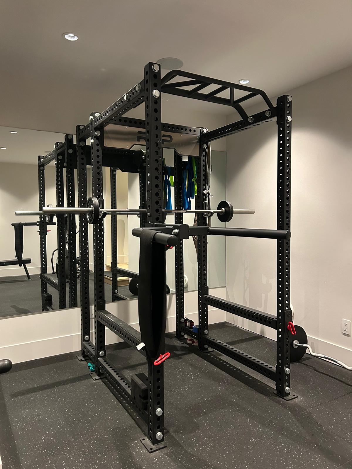 Rogue power rack with loaded barbell, safety arms, hanging resistance bands, and adjustable bench inside, in front of a full mirror wall in the Abbotsford home gym build.