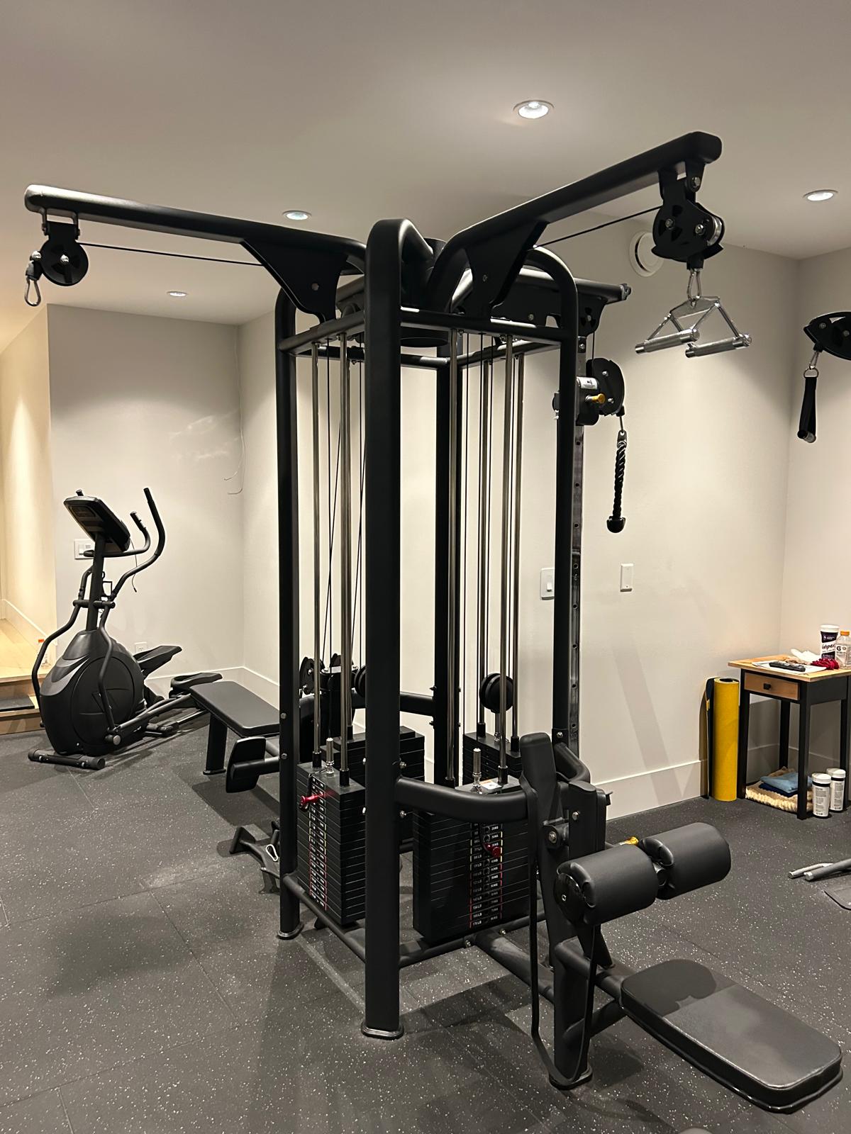 Dual-pulley functional cable station with leg extension attachment beside a touchscreen elliptical in the Abbotsford home gym build.