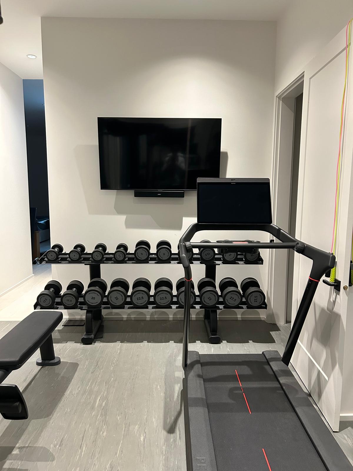 Cardio and media wall with mounted TV, Bose soundbar, dumbbell rack, and touchscreen treadmill in the Kelowna home gym.